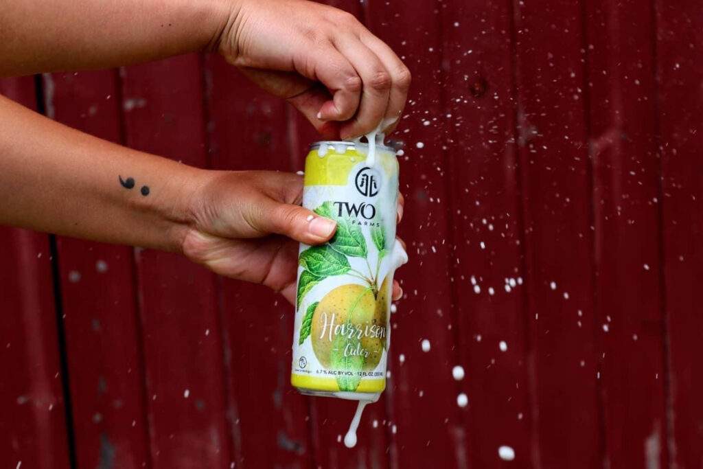 A photo of a person's hands opening a can of Two K Farms cider in front of a red wood background.
