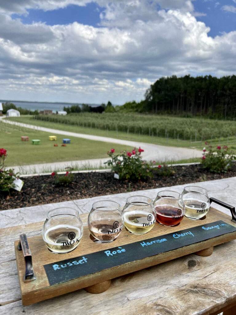 Photo of a flight of cider taken from the Two K Farms tasting room patio. Photo taken on a summer day overlooking the orchard and lawn games in the background.