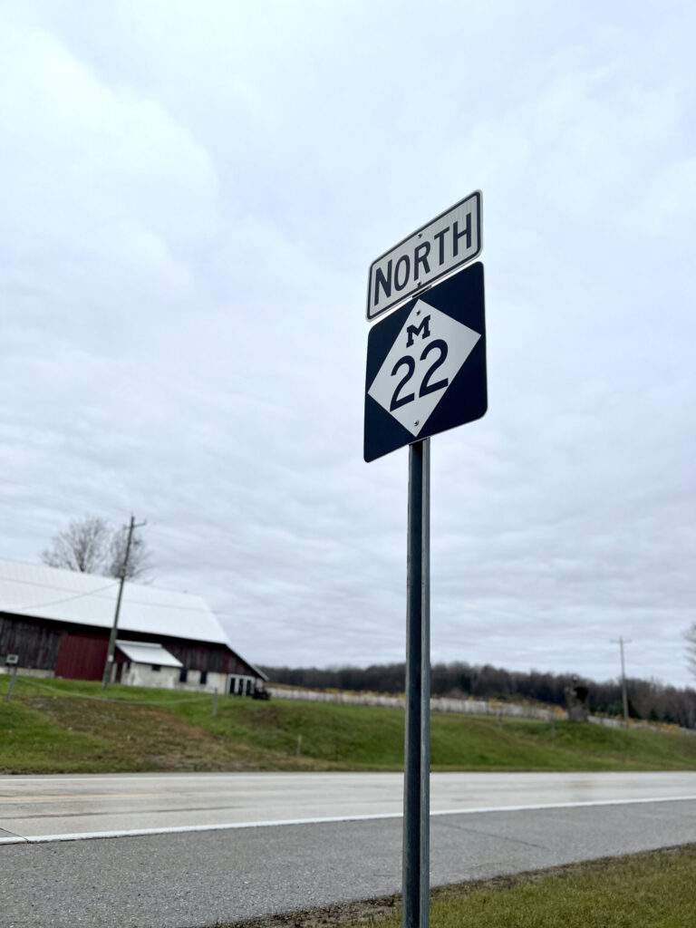 Photo of the North M-22 Road sign next to the roadway, with overcast sky and a barn in the background.