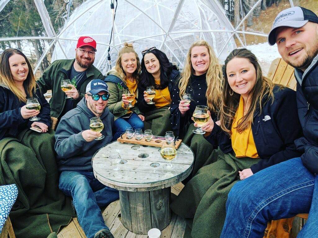 Group of friends smiling while holding Two K Farms Cider glasses. They are sitting under an igloo tent on the Two K Farms patio.