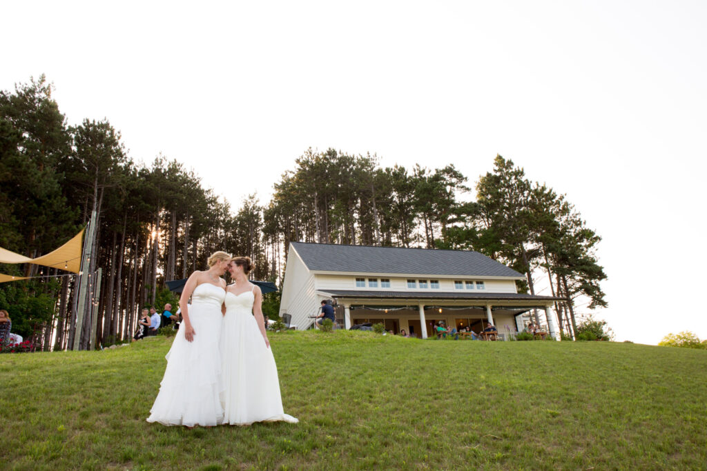 Photo of two women in wedding dresses standing in front of the Two K Farms Tasting Room with a sunset in the background.
