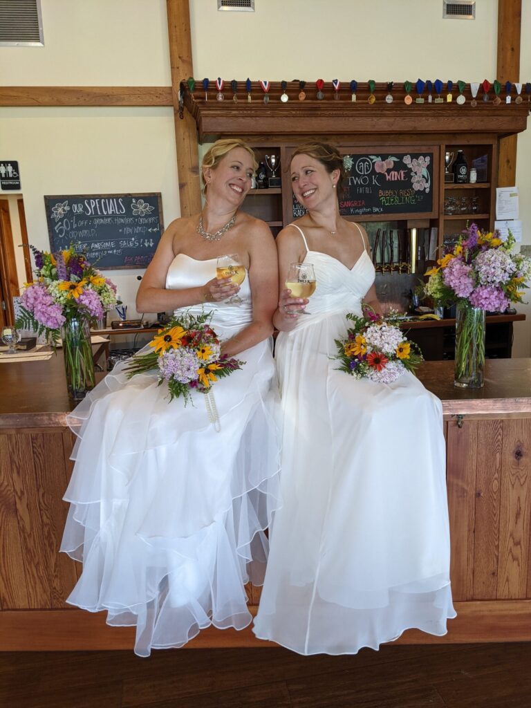 Photo of two brides sitting on the tasting room bar holding Two K Farms wine glasses and bouquets of flowers.