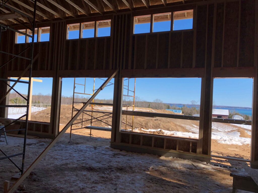 Photo taken from the inside of a newly constructed wooden building. The photo looks out over the construction site with a barn and water in the background.