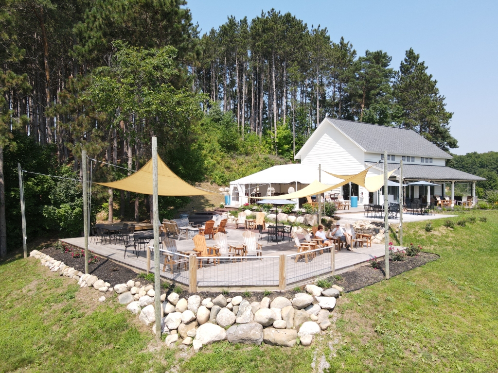 Photo of the tasting room, patio, and deck on a sunny summer day. There are yellow canopies hanging over the deck with pine trees in the background and green grass in the foreground.