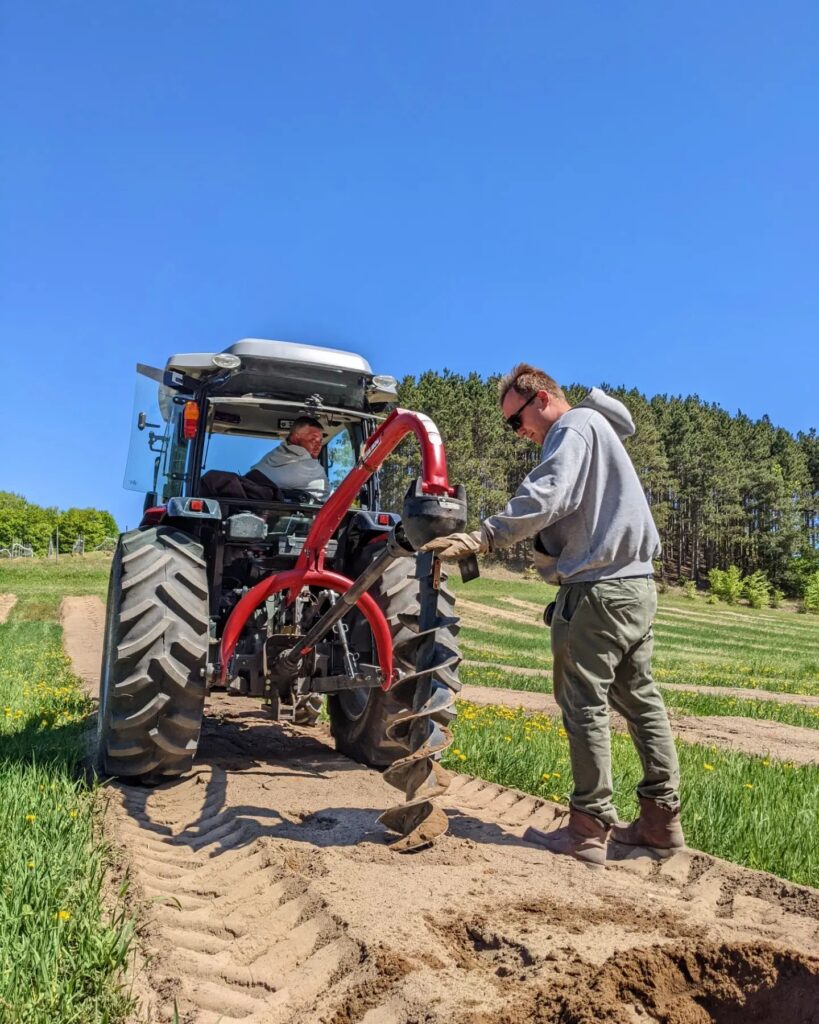 Photo of a tractor in the field of an orchard that is equipped with an auger on the back. A man is in the tractor looking back at another man who is operating the auger.