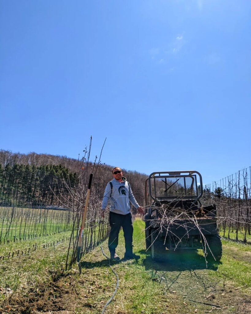 Photo of man standing between rows of newly planted apple trees in an apple orchard smiling at the camera next to an ORV, with trees and blue sky in the background.