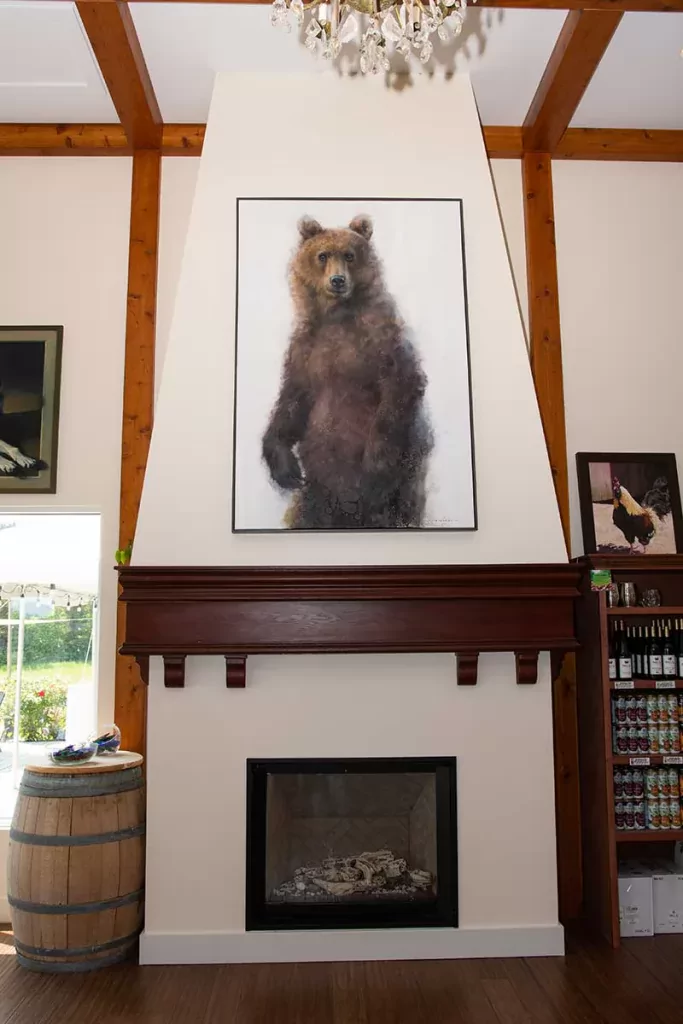 Photo of the interior of the Two K Farms tasting room. There is a fireplace with a wooden mantel, and above that a large panting of a bear is handing from the wall. There is a wine barrel to the left of the fireplace and shelves that hold Two K Farms cider cans to the right.