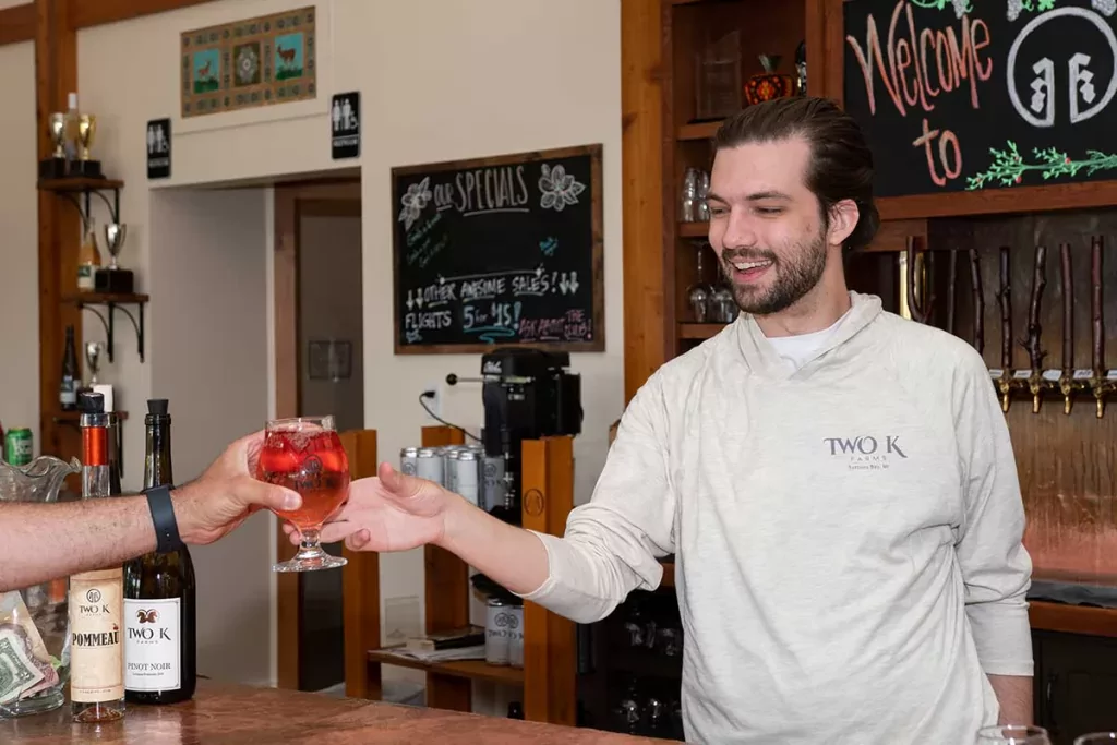 Photo of Cooper from the Two K Tasting Room staff handing a glass of rose cider over the bar of the Two K Farms tasting room to a person who is out of frame.