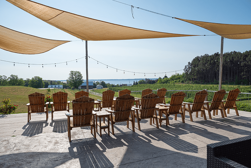 Photo of the Two K Farms Tasting Room patio with several wooden chairs facing the green orchard and vineyard that overlooks Grand Traverse Bay. There is water in the background and blue sky.