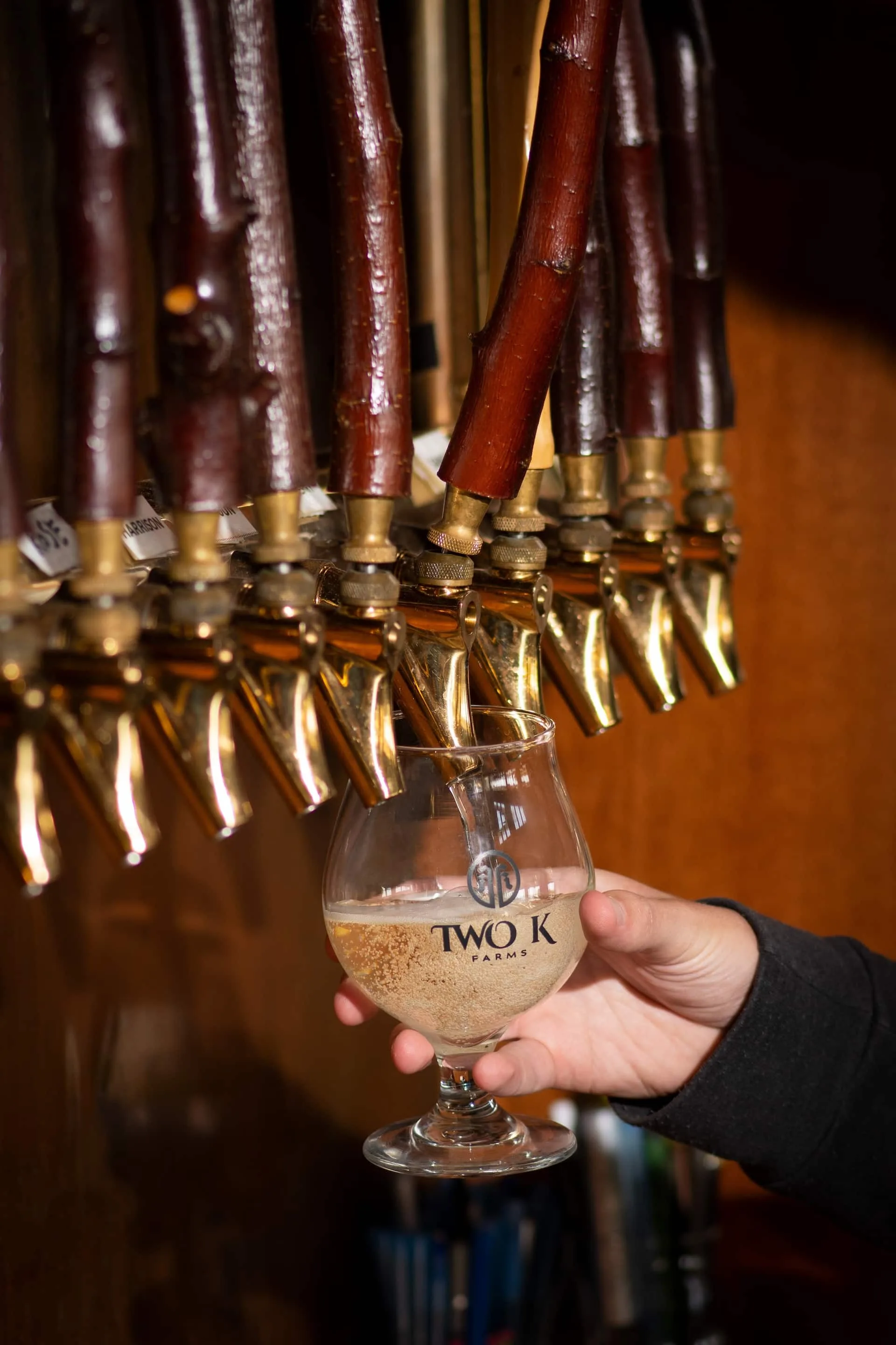 Hand holding a goblet style glass with the Two K Farms logo under a row of cider taps in the Two K Farms tasting room. One tap has been pulled and is pouring into the glass