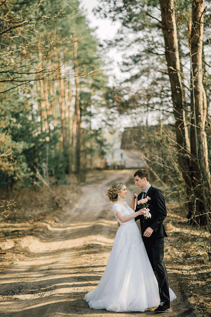 Photo of a bride and groom standing in a wooded area on a gravel driveway. A white building is behind them in the distance.